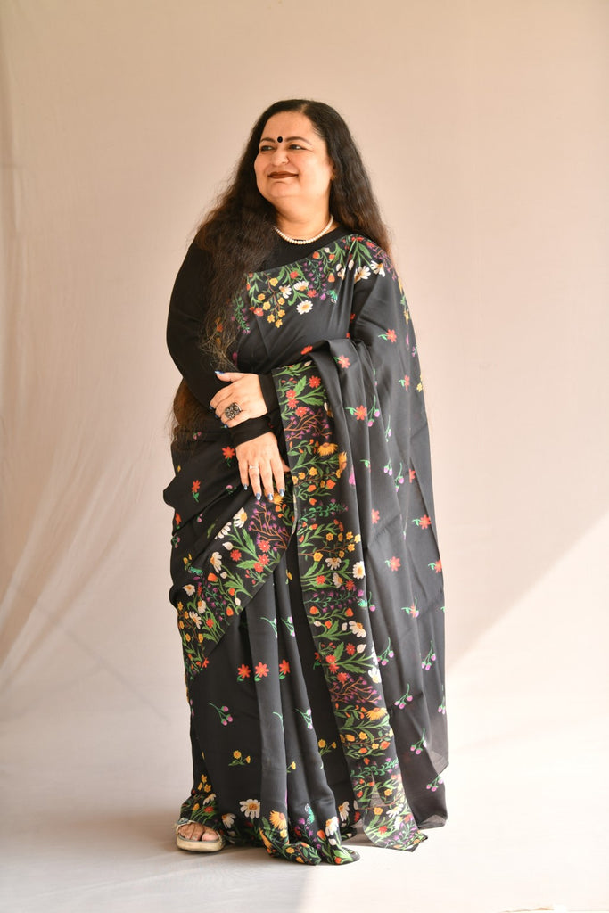 Female model wearing black designer floral print saree in front post with black blouse.